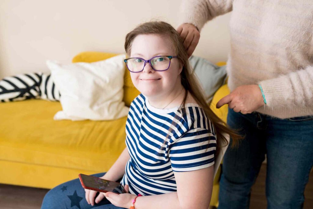 Ein junges Mädchen mit Down-Syndrom sitzt auf einem Stuhl und spielt konzentriert ein Spiel auf dem Smartphone, während ihr im Hintergrund liebevoll die Haare frisiert werden – ein Moment digitaler Teilhabe und Freude am mobilen Gaming.