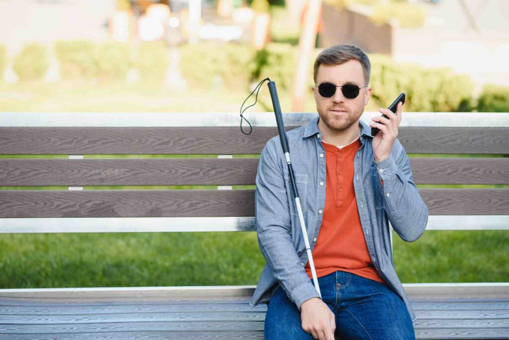 Ein blinder Mann sitzt auf einer Parkbank, hält ein Smartphone vor sich und nutzt es per Sprachsteuerung. In der anderen Hand hält er einen weißen Langstock – ein Beispiel für digitale Barrierefreiheit im Alltag durch sprachgesteuerte Webnavigation.